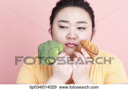 Stock Photo - a chubby woman holding broccoli and cupcake. Fotosearch