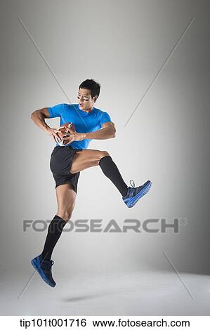 Stock Photograph - a fit man in blue shirt jumping with rugby ball. Fotosearch