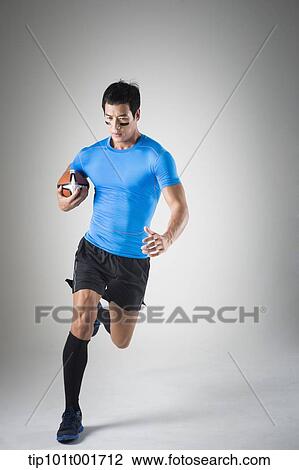 Stock Image - a fit man in blue shirt running with rugby ball. Fotosearch