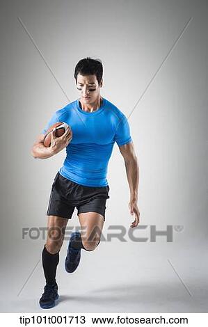 Stock Image - a fit man in blue shirt running with rugby ball. Fotosearch
