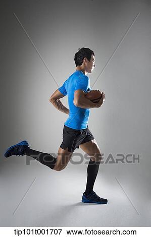 Stock Photo - a fit man in blue shirt running with rugby ball. Fotosearch