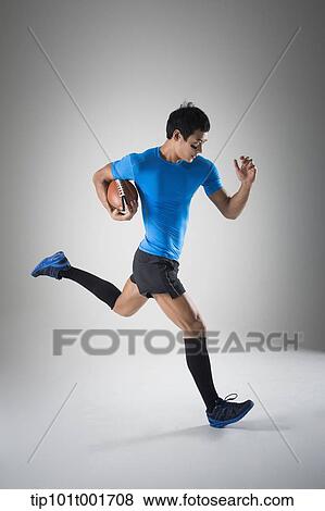 Stock Photo - a fit man in blue shirt running with rugby ball. Fotosearch