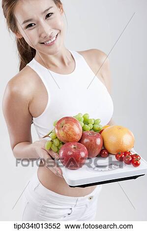 Stock Image - a healthy looking woman holding fruits on weighing scale. Fotosearch