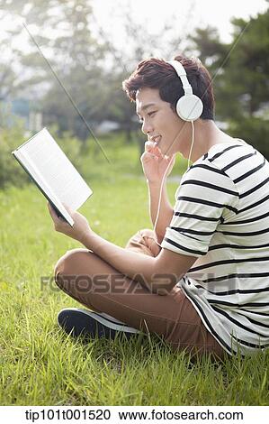Stock Image - a man sitting on the grass listening to music while reading a book. Fotosearch
