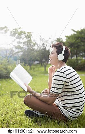 Stock Image - a man sitting on the grass listening to music while reading a book. Fotosearch