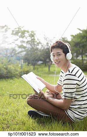Stock Image - a man sitting on the grass listening to music while reading a book. Fotosearch