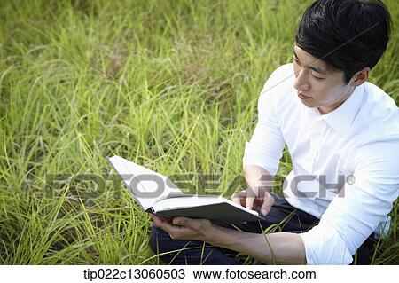 Stock Image - a man sitting on the grass reading a book. Fotosearch