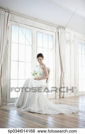 Stock Photo - a woman sitting on a chair with wedding dress and flower bouquet . Fotosearch