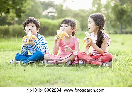 Picture - kids sitting on the grass eating bananas. Fotosearch