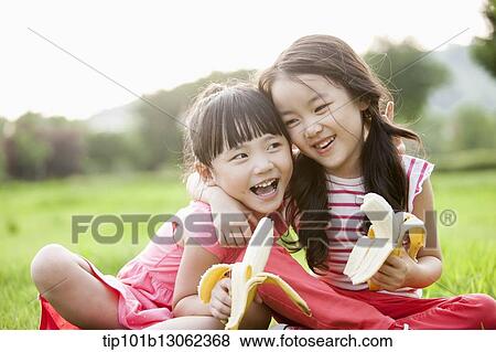 Stock Photo - two girls sitting on grass eating bananas. Fotosearch