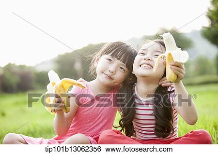 Stock Photograph - two girls sitting on grass eating bananas. Fotosearch