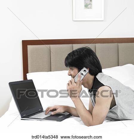 Stock Photo - a man talking on a phone and looking at a notebook on a bed. Fotosearch