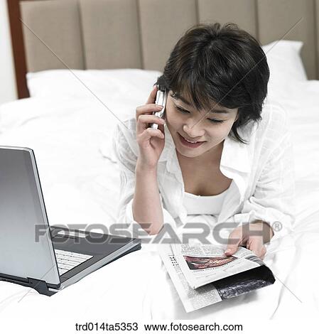Stock Image - a woman talking on a phone and looking at a notebook on a bed. Fotosearch