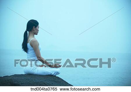 Stock Photography - a woman doing Yoga on the rock in a seashore. Fotosearch