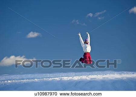 Stock Image - Young woman jumping in snow, arms outstretched. Fotosearch