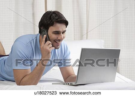 Stock Image - Man talking on the phone while working on a laptop. Fotosearch