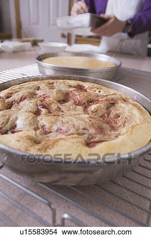 Picture - baked cake in a round pan on the cooling rack with baker in the background. Fotosearch