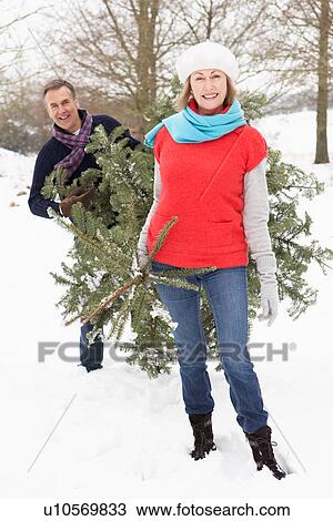 Stock Image - Senior Couple Carrying Christmas Tree In Snowy Landscape. Fotosearch
