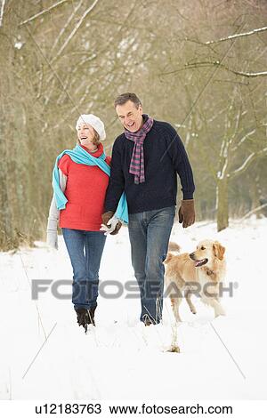 Stock Image - Senior Couple Walking Dog Through Snowy Woodland. Fotosearch