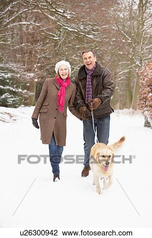 Stock Image - Senior Couple Walking Dog Through Snowy Woodland. Fotosearch