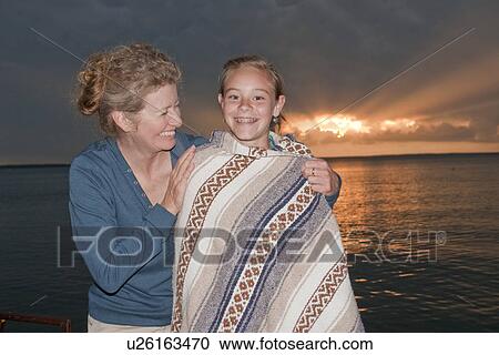 Stock Image - 50-s mom with teen wrapped in blanket by Clear Lake at sunset, Riding Mountain National Park, Manitoba, Canada. Fotosearch