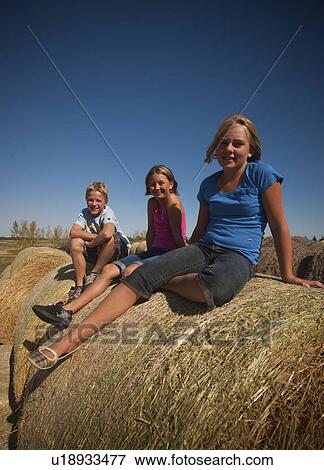 Stock Photo - Boy (12) girl (10) and girl (12) seated on bales of straw; Redvers, Saskatchewan, Canada. Fotosearch