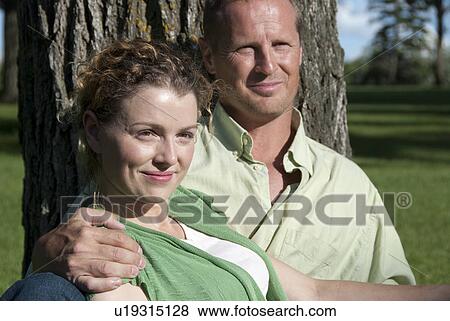 Stock Photo - Couple in their 40's seated at base of tree in park, Winnipeg, Canada. Fotosearch