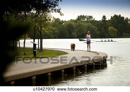 Stock Image - Female runner lakeside in park with dog, Regina, Saskatchewan. Fotosearch