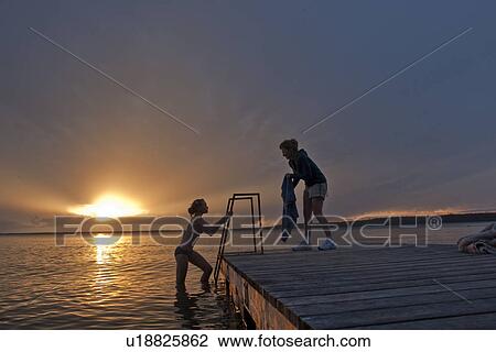 Stock Image - Girl climbing out of water onto dock, mom holding towel; Riding Mountain National Park, Manitoba, Canada. Fotosearch