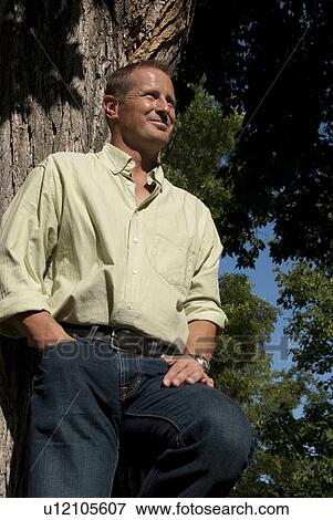 Stock Photo - Low angle view of man in 40's leaning against tree, Winnipeg, Canada. Fotosearch