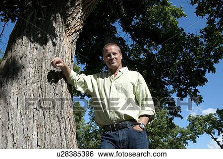 Stock Photograph - Lower angle view man in 40's leaning on tree in park, Wnnipeg, Canada. Fotosearch