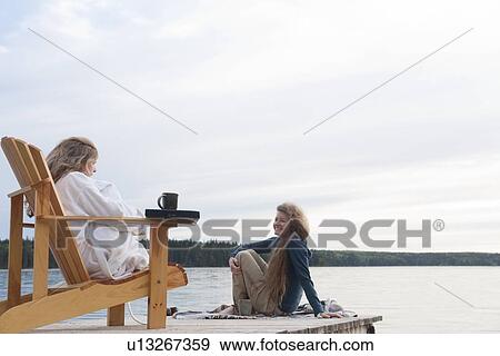 Stock Photo - One woman seated in deck chair, second woman seated on dock with coffee, Clear Lake, Manitoba, Canada. Fotosearch