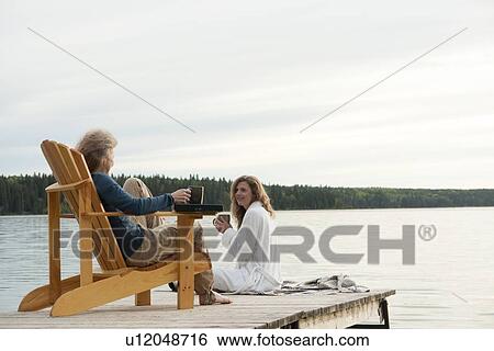 Stock Photograph - One woman seated in deck chair, second woman seated on dock with coffee, Clear Lake, Manitoba, Canada. Fotosearch