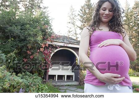 Picture - Pregnant woman holding abdomen in front of garden gazebo, Manitoba, Canada. Fotosearch