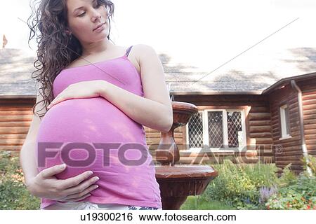 Stock Photograph - Pregnant woman holding abdomen in front of garden fountain, Manitoba, Canada. Fotosearch