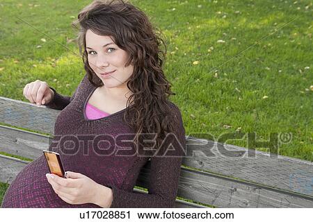 Stock Image - Pregnant woman on park bench holding cell phone, Manitoba, Canada. Fotosearch