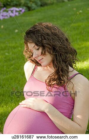 Stock Photo - Pregnant woman seated on grass with hand on abdomen, Manitoba, Canada. Fotosearch