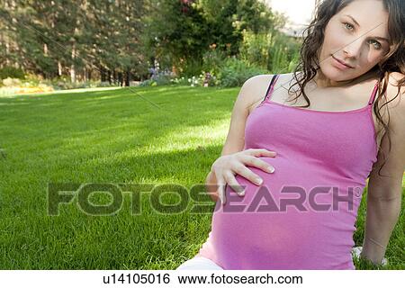 Stock Photograph - Pregnant woman seated on grass with hand on abdomen, Manitoba, Canada. Fotosearch