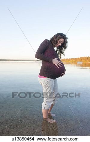 Picture - Pregnant woman with hands on abdomen wading in Clear Lake, Manitoba, Canada. Fotosearch