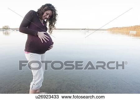 Stock Image - Pregnant woman with hands on abdomen wading in Clear Lake, Manitoba, Canada. Fotosearch