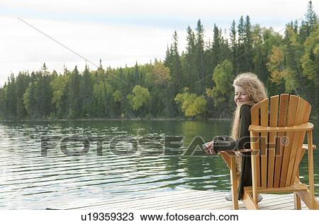 Stock Image - Shoreline of trees with woman seated in deck chair on dock, Clear Lake, Manitoba, Canada. Fotosearch