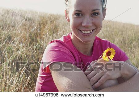Stock Photograph - Smiling woman holding small flower on hillside, rural Manitoba, Canada. Fotosearch