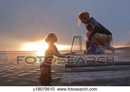 Stock Image - Tween girl in water, mid-50's woman seated on dock, sunset, Riding Mountain National Park, Manitoba, Canada. Fotosearch