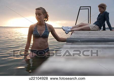 Stock Photo - Tween girl wading in water, mom seated on dock, Riding Mountain National Park, Manitoba, Canada. Fotosearch