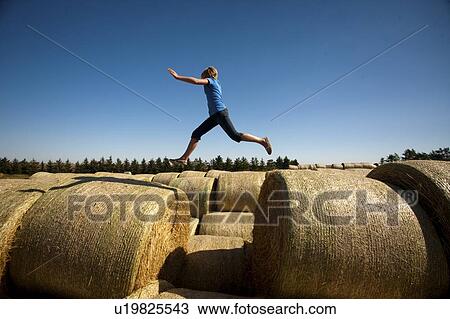 Stock Image - Twelve year old girl leaps between bales; Redvers, Saskatchewan, Canada. Fotosearch