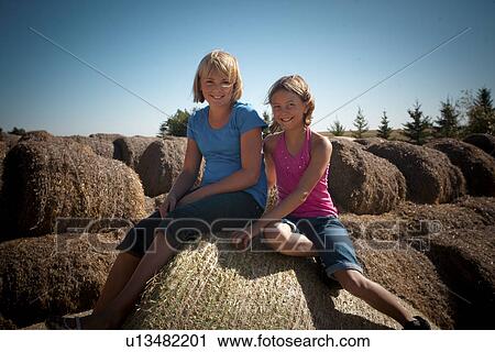 Stock Image - Two girls, 10 and 12, seat on bales of straw; Redvers, Saskatchewan, Canada. Fotosearch