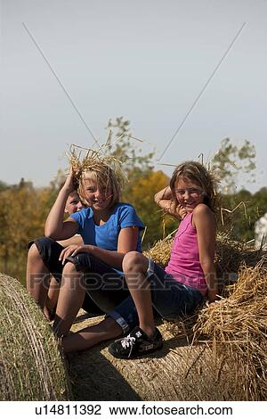 Stock Image - Two girls (12 and 10) holding staw as if it were hair, seated on bales; Redvers, Saskatchewan, Canada. Fotosearch