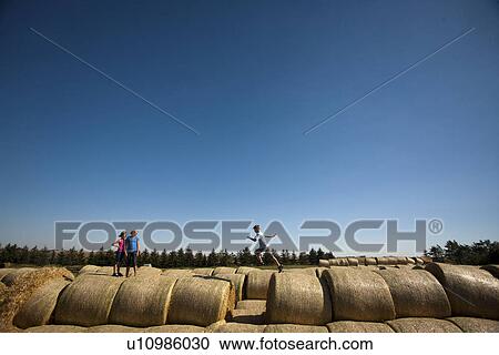 Stock Image - Two girls watch boy jump over space between bales; Redvers, Saskatchewan, Canada. Fotosearch
