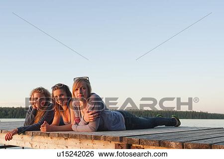 Stock Photograph - Two teen girls and woman in 50's lying on dock by lake, Riding Mountain National Park, Manitoba, Canada. Fotosearch