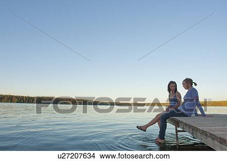 Picture - Two teen girls seated on dock of Clear Lake, Riding Mountain National Park, Manitoba, Canada. Fotosearch